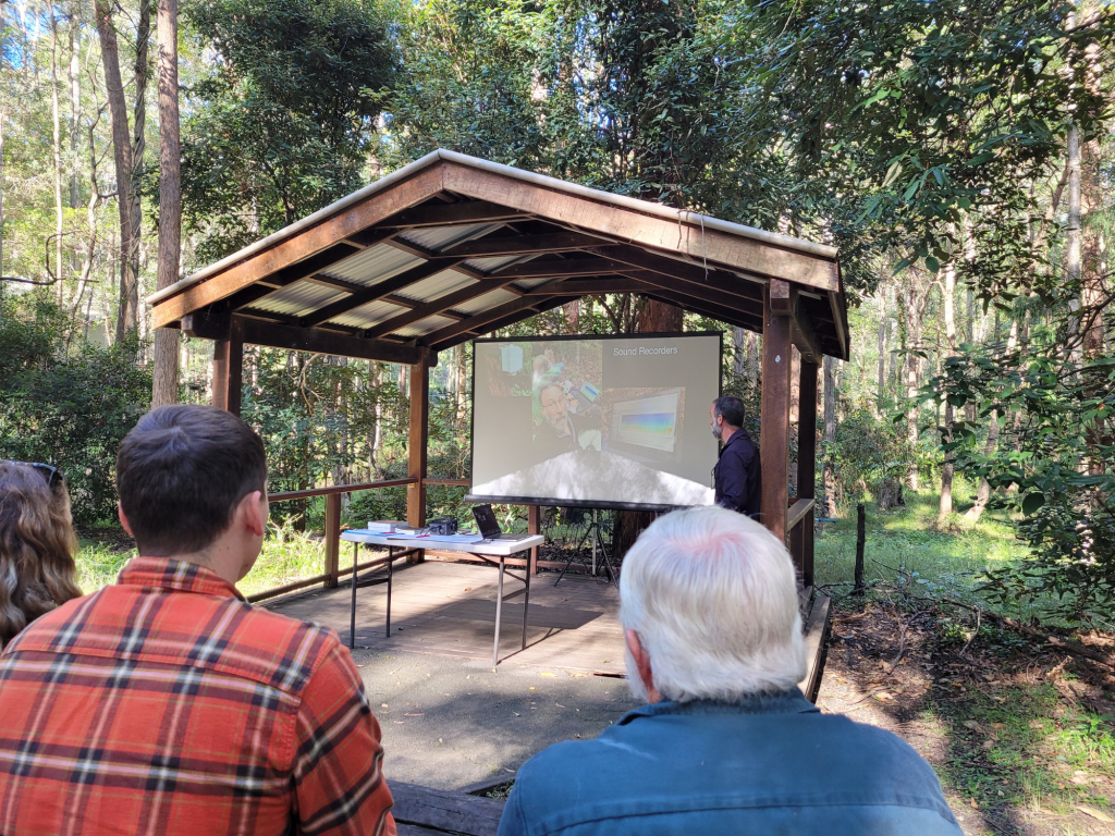 Outdoor presentation in a bushland setting, with a speaker discussing sound recorders to an audience seated under a thatched roof.