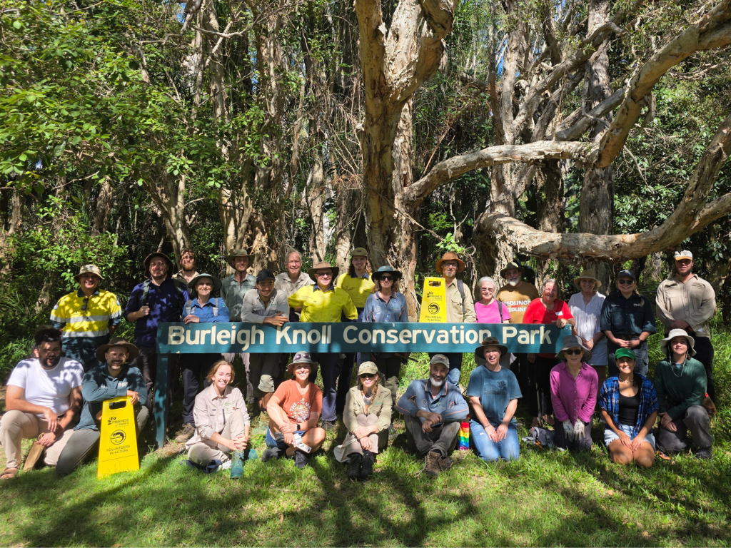 A group of volunteers posing in front of a sign for Burleigh Knoll Conservation Park, surrounded by trees and greenery on the Gold Coast.