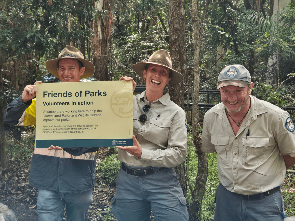 Three volunteers from the Friends of Parks program smiling while holding a sign that reads 'Friends of Parks, Volunteers in Action' in a natural forest setting.