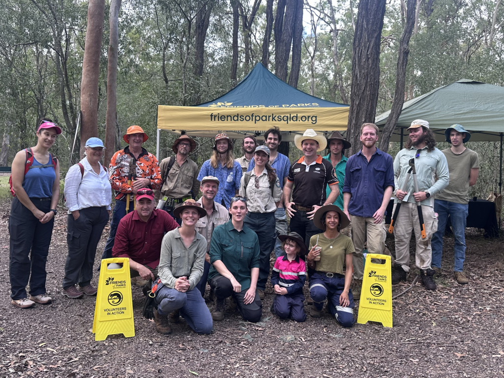 Group of volunteers standing in a forest area, posing in front of a banner for 'Friends of Parks Queensland'. They are wearing various outdoor clothing and hats, with a sign reading 'Volunteers in Action' in the foreground.