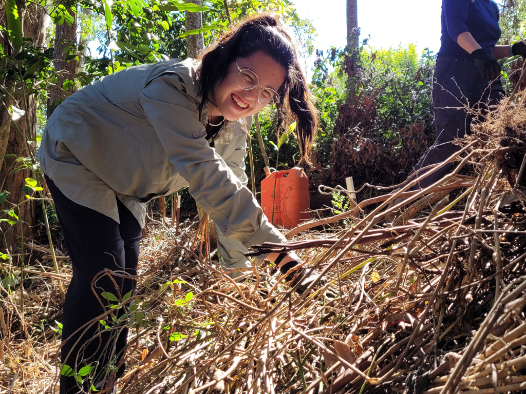 A smiling woman with glasses kneeling in a green bushy area, working among dry twigs and plants. She is wearing a light jacket and focused on her task, with another person visible in the background.