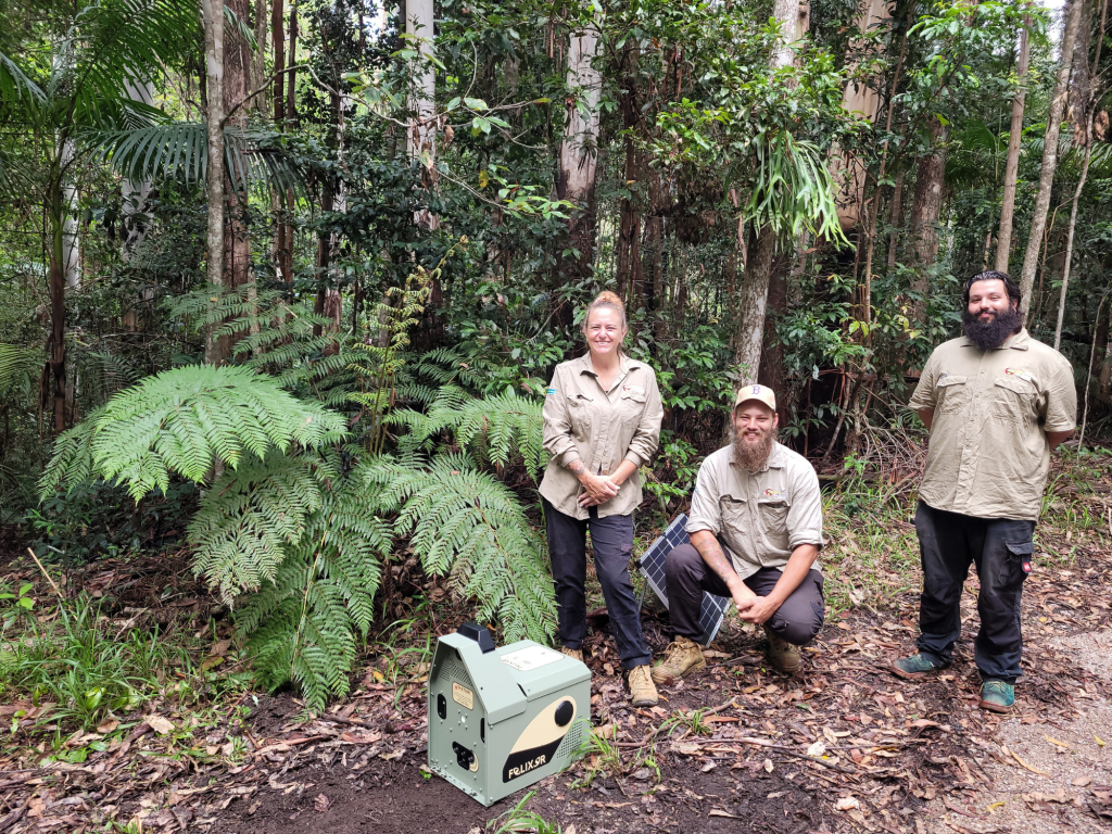 Three individuals stand in a lush forested area surrounded by trees and ferns, with a generator placed on the ground.