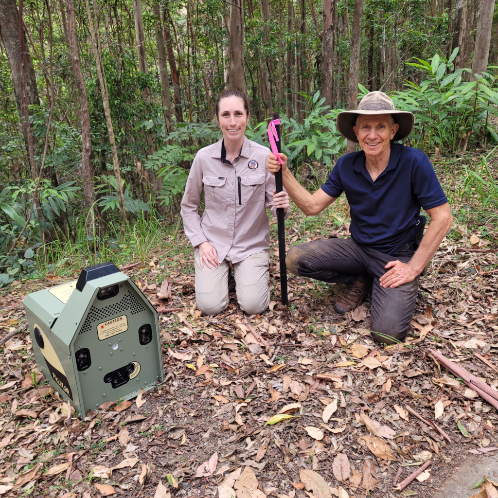 Two individuals kneeling beside a wildlife monitoring device in a forested area, surrounded by green foliage and fallen leaves.